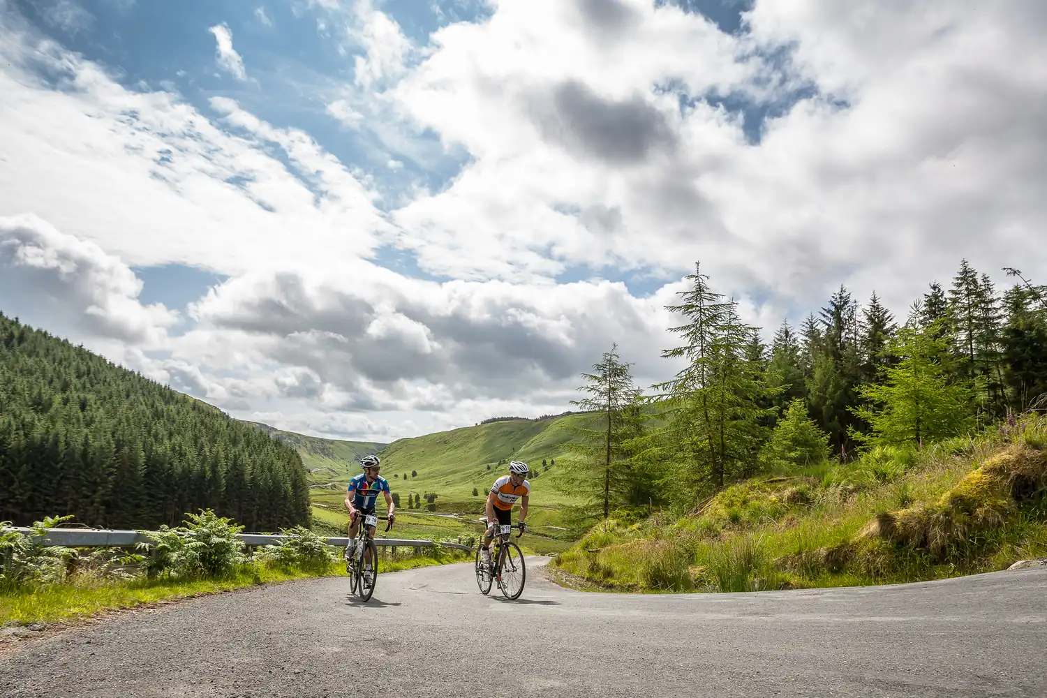 Two cyclists climbing a forested Welsh valley road under a big sky